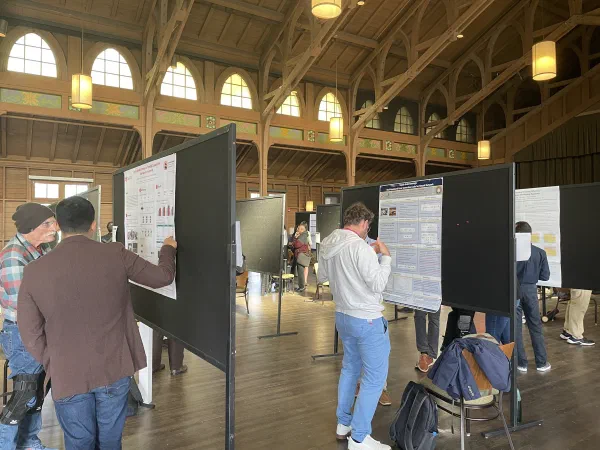 Researchers gather around poster boards in a sunlit wooden hall.